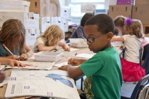 Children in an elementary school classroom using pens and pencils to complete work in a workbook.